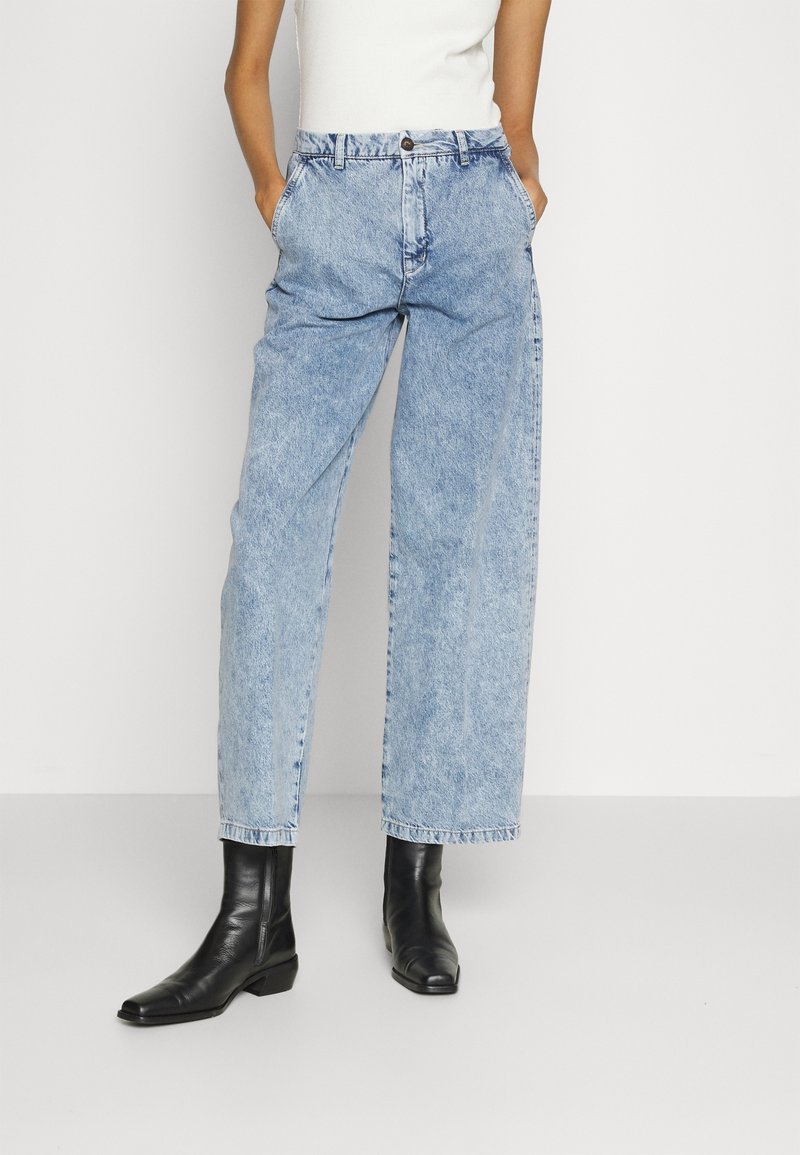Personne portant un jean bleu clair taille haute à jambes larges, un haut blanc sans manches et des bottines en cuir noir, debout les mains dans les poches.