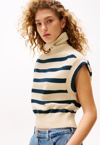 Gestrickter kurzärmeliger Pullover in Creme mit marineblauen horizontalen Streifen, mit hohem Kragen und geripptem Saum; kombiniert mit hellblauen Jeans.