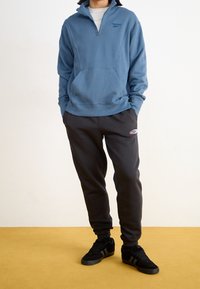 Blå fleecetröja med kvartszip och framficka, bärs över en grå t-shirt, kombinerad med svarta mjukisbyxor med logotypmärke och svarta sneakers.