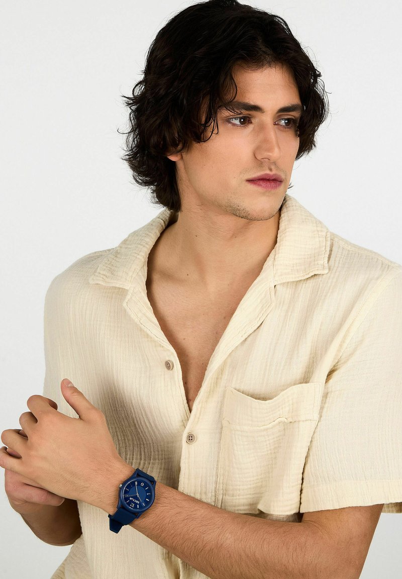 Orologio in silicone blu con quadrante rotondo, indicatori e lancette bianche. Modello che indossa una camicia crema con texture, mostrando l'orologio sul polso sinistro.