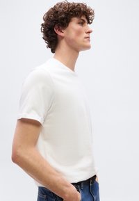 Νέος άνδρας με σγουρά μαλλιά που φοράει λευκό απλό t-shirt και μπλε τζιν, στέκεται στο πλάι με τα χέρια στις τσέπες μπροστά σε λευκό φόντο.