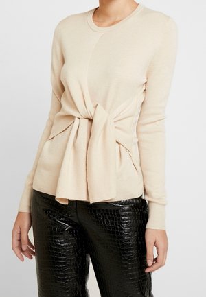 Haut beige à manches longues avec un détail de lien à l'avant, associé à un pantalon noir en similicuir avec une texture crocodile.