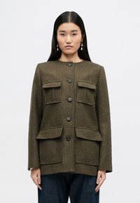 Brązowa marynarka w kratę z okrągłym dekoltem, zamknięciem na guziki z przodu i dwiema dużymi kieszeniami na piersiach; cechuje się dopasowanym krojem i długimi rękawami.