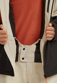 Geïsoleerde jas met een beige buitenkant, zwarte accenten en een ritszak. Gelaagd met een textuur, oranje fleece eronder.