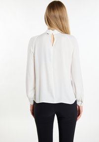 Weiße Bluse mit langen Ärmeln, glatter Textur, Knopfverschluss am Rücken mit Schlüssellochdetail, kombiniert mit schwarzen, eng anliegenden Hosen.