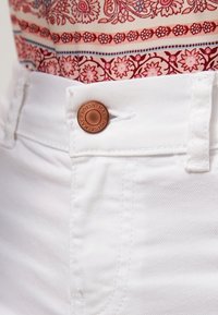 Valkoiset farkut, joissa on pronssin värinen nappi, johon on kaiverrettu teksti. Kankaan pinta on sileä, ja saumoissa on näkyvissä ompeleet.