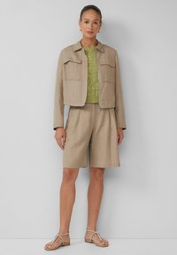 Veste beige courte avec deux poches avant, top vert avec un motif géométrique doré, shorts plissés beiges et sandales argentées.