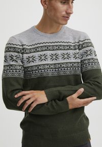 Mørkegrøn strikket sweater med en grå mønstret bærestykke. Designet har snefnugs- og geometriske mønstre i hvid, mørkegrøn og sort. Ribbekanter ved håndleddene.