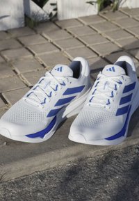 White athletic shoes with blue accents, lightweight mesh upper, textured surface, padded collar, and lacing system with prominent branding.