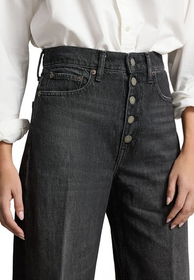 Augsta vidukļa melni džinsi ar pogu aizdari, piecām kabatām un nedaudz teksturētu virsmu. Apģērbs kombinēts ar baltu krekli ar pogām.