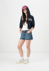 Tamsiai mėlyna trumpa sweatshirtytė su užrašu "UNIVERSITY", balta marškinėliai be rankovių, džinsinė mini sijonas ir šviesūs sportbačiai su kojinėmis. Raudona beisbolo kepuraitė suteikia kontrastą.