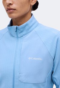 Γυναίκα που φοράει ένα ανοιχτό μπλε ζακέτα Columbia με φερμουάρ, υφή υφάσματος και ψηλό γιακά, εμφανίζοντας το λογότυπο της μάρκας στο στήθος.
