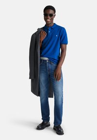 Muž v slunečních brýlích, modrém polo tričku, džínách a černých botách, držící šedý kabát přes jedno rameno, usmívající se na bílém pozadí.