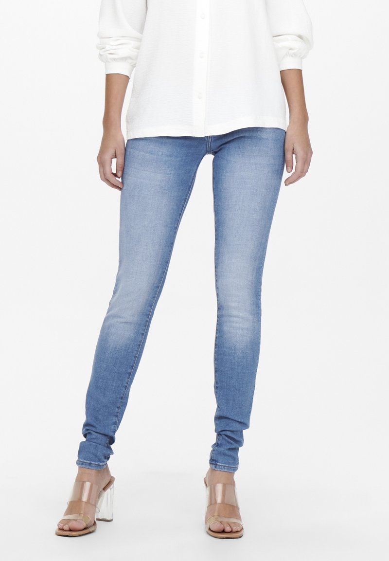 Femme portant un jean skinny bleu clair, une chemise blanche à boutons aux manches retroussées et des sandales à talons hauts avec des brides transparentes.