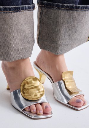 Pies con zuecos plateados de tacón alto y punta abierta con grandes adornos dorados decorativos, combinados con vaqueros de mezclilla remangados.