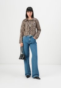 Kvinna står klädd i en jacka med leopardsmönster, blå vida jeans, svarta spetsiga skor och håller en svart handväska med ena handen i fickan.
