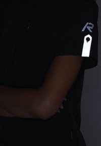 Personne les bras croisés portant une chemise noire avec un logo bleu "R" et un onglet blanc réfléchissant sur la manche.