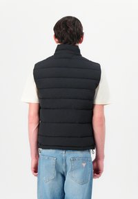 Vest negru cu umplutură, cu guler înalt, design cusut și tiv cu șnur, purtat peste o cămașă cu mânecă scurtă de culoare deschisă și blugi.