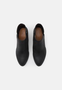 Chaussures en cuir noir avec une texture tissée, bout pointu et fermetures éclair sur les côtés. Doublure intérieure marron visible. Semelle plate.
