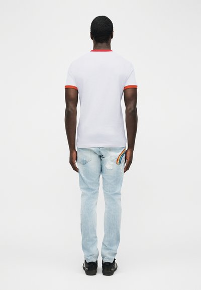 Άνδρας γυρισμένος πλάτη που φοράει λευκό μπλουζάκι με κόκκινη επένδυση στα μανίκια και το γιακά, ανοιχτόχρωμο μπλε τζιν με κέντημα ουράνιου τόξου στην πίσω τσέπη και μαύρα παπούτσια.