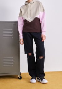 Huvtröja i beige, rosa och brunt med en geometrisk färgblockdesign. Tillsammans med svarta slitna jeans och sneakers.