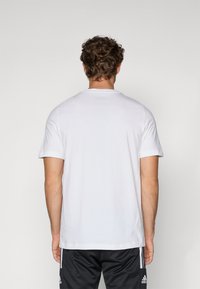 T-shirt blanc à col rond fabriqué en coton, avec des manches courtes et une coupe droite. Le tissu semble lisse avec une texture douce.