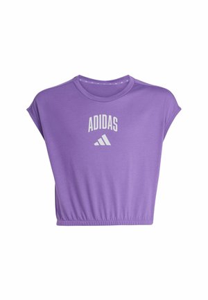 Persona joven con ropa deportiva púrpura de Adidas sosteniendo una botella de agua negra, de pie con zapatos blancos sobre un fondo liso.