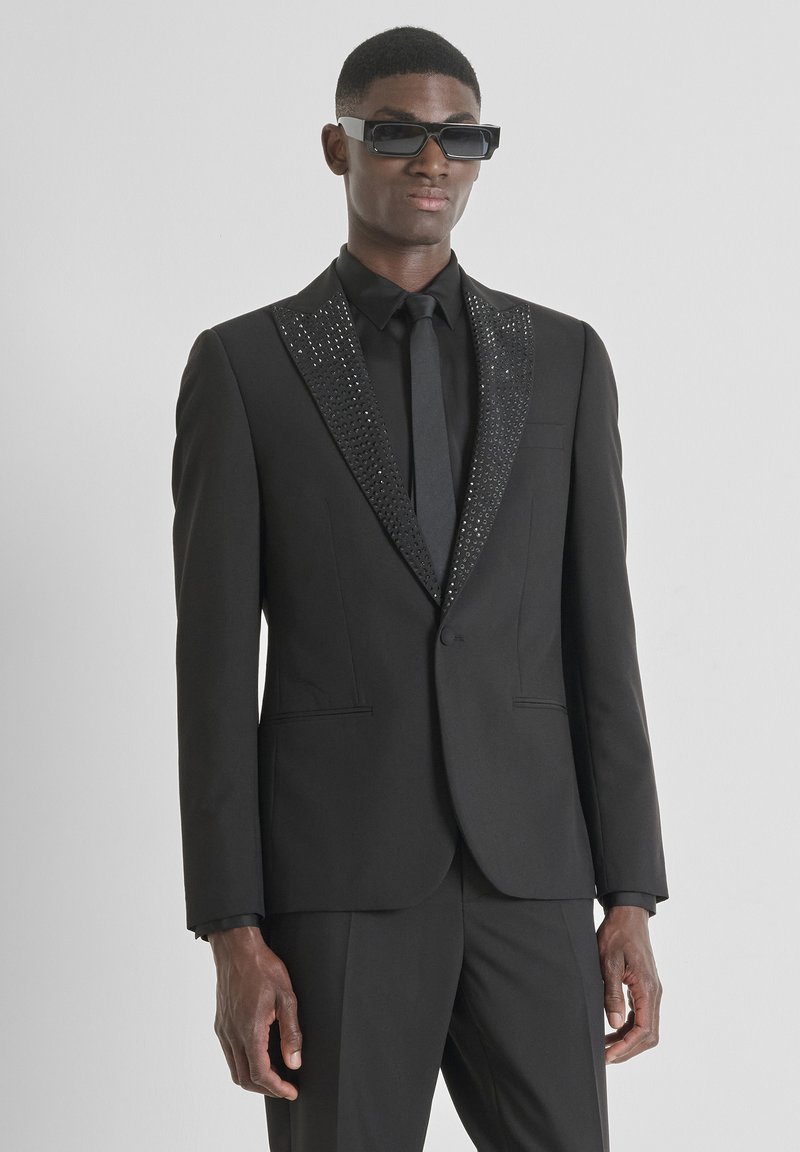 Zwart smoking met een gestructureerde, fonkelende zwarte pailletten revers, strak ontwerp en bijpassende stropdas. Draagt bijpassende minimalistische zwarte zonnebril.