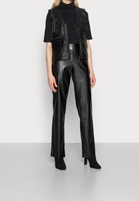 Chaleco de cuero negro con múltiples bolsillos, combinado con pantalones de cuero negro de cintura alta. El modelo lleva botines negros de tacón.