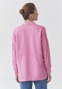 Blusa rosa a maniche lunghe con colletto, caratterizzata da una vestibilità ampia e spalle scese, abbinata a jeans azzurri chiari. Tessuto morbido.