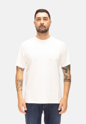 Uomo con capelli corti e scuri e barba, indossa una semplice maglietta bianca e jeans scuri, in piedi su uno sfondo grigio chiaro.