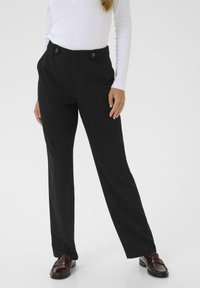 Pantaloni neri sartoriali con gamba dritta, caratterizzati da pince frontali e due bottoni decorativi in vita. Indossati con una maglietta bianca a maniche lunghe.