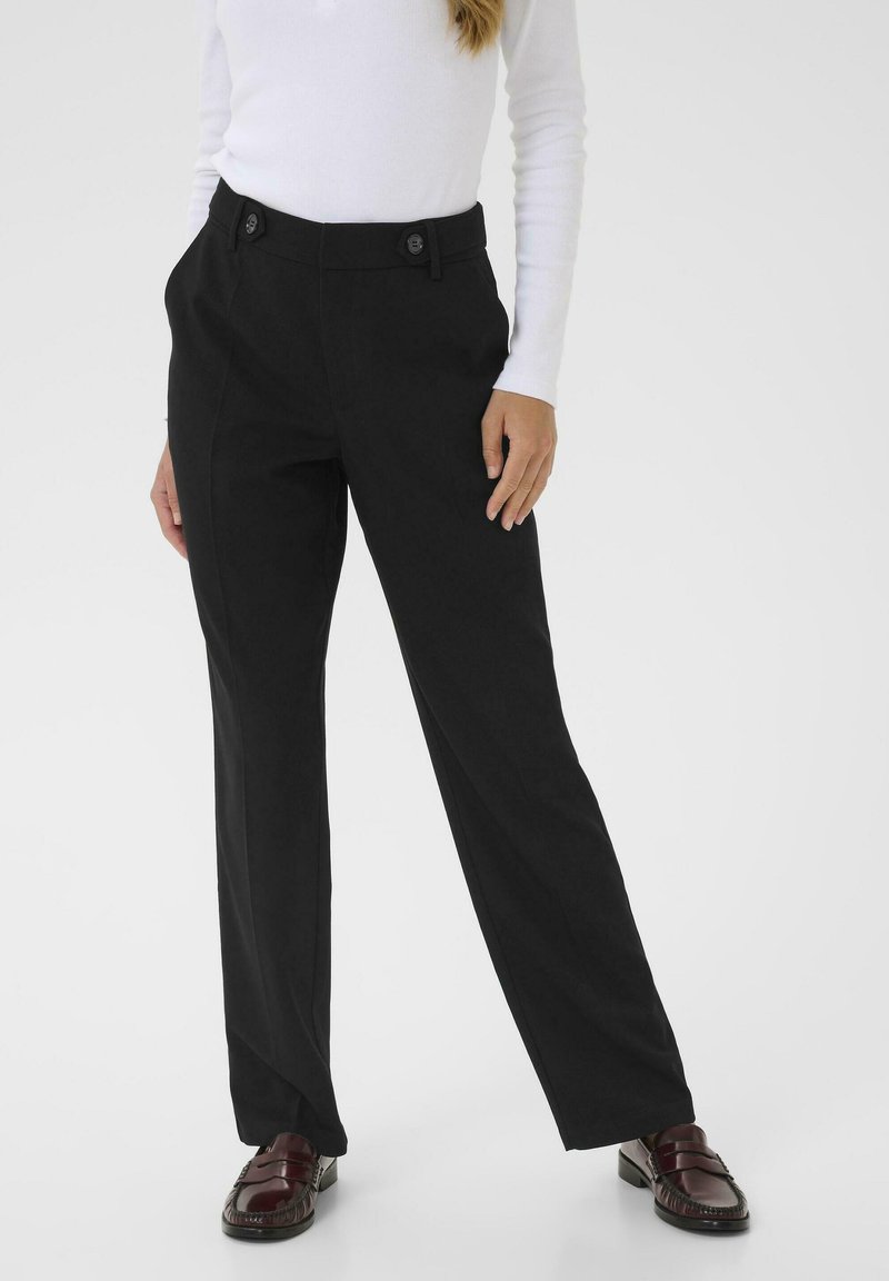 Pantaloni neri sartoriali con gamba dritta, caratterizzati da pince frontali e due bottoni decorativi in vita. Indossati con una maglietta bianca a maniche lunghe.