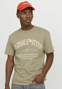 Olijfgroene katoenen T-shirt met witte tekst en logo-ontwerp; korte mouwen en ronde hals. Gecombineerd met een rode pet met witte tekst.
