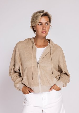 Frau mit kurzen blonden Haaren, die einen beigen Kapuzenpullover mit Reißverschluss und eine weiße Hose trägt, steht mit den Händen in den Taschen vor einem einfarbigen Hintergrund.