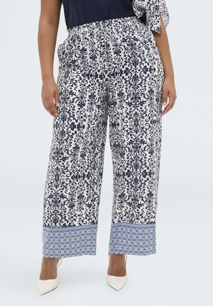 Kobieta w białych i granatowych, wzorzystych spodniach o szerokich nogawkach z elastycznym pasem oraz białych, szpiczastych szpilkach, stojąca na jednolitym tle.