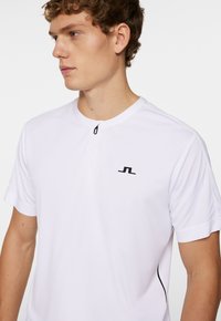 Polo shirt bianco a maniche corte con una mezza abbottonatura, logo nero sul petto e sottili dettagli laterali neri. Tessuto dalla texture liscia.