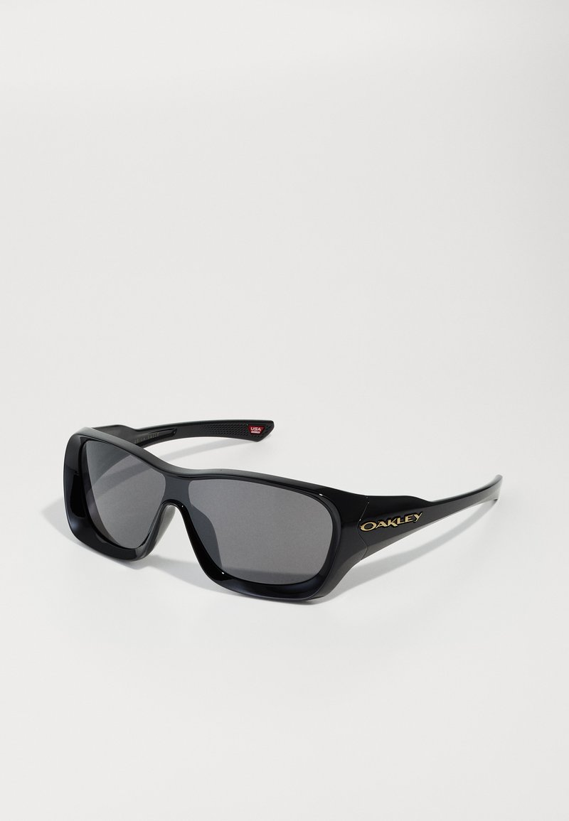Black sunglasses with a wide rectangular frame, dark gray lenses, and gold Oakley logo on the side. High-gloss plastic material.