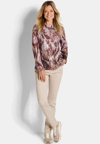 Langärmliges Oberteil im burgunder- und braunen Batikmuster mit Paillettenakzenten. Kombiniert mit hellbeigen Slim-Fit-Hosen und neutralen Sneakers.