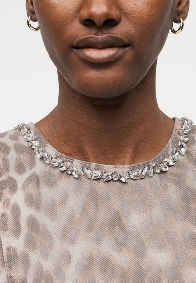 Κοντινό πλάνο ενός ατόμου που φορά χρυσούς κρίκους στα αυτιά και ένα μπεζ τοπ με σχέδιο λεοπάρδαλης και λαιμόκοψη διακοσμημένη με καθαρές κρυστάλλινες διακοσμήσεις.