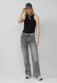 Schwarzes ärmelloses Oberteil, graue weite Jeans, schwarze Mütze mit Logo und gemusterter Schal. Das Modell steht vor einem schlichten Hintergrund.