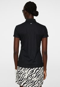 Czarna koszulka polo o fakturowanym wykończeniu, z krótkimi rękawami i kołnierzykiem. Widok z tyłu pokazuje białe logo blisko dekoltu. Połączona z spódnicą w zebrze.
