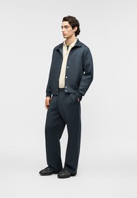 Chaqueta azul marino oscuro con cierre de botones, combinada con una camisa amarilla clara y pantalones azul marino de corte holgado. Zapatillas negras completan el conjunto.