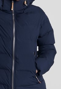 Tmavě modrá péřová bunda se zipem, kapucí a bočními kapsami. Látka je hladká, s prošívanou texturou a kovovými detaily.