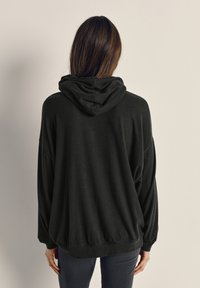 Osoba s dlhými hnedými vlasmi, oblečená v čiernej mikine s kapucňou a tmavých nohaviciach, viditeľná zozadu na jednoduchom pozadí.