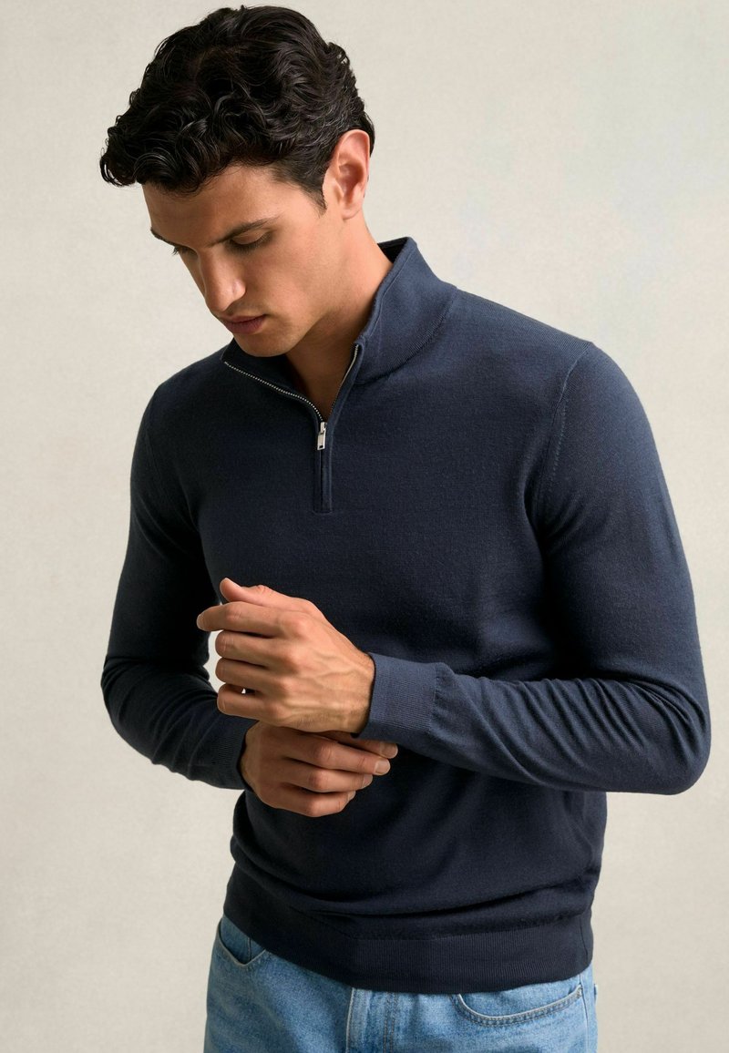 Marineblauer Reißverschluss-Pullover aus einem glatten, dehnbaren Stoff. Enganliegende lange Ärmel und gerippte Bündchen, mit einem halbseitigen Reißverschluss am Hals und minimalistischem Design.