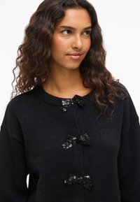 Svart stickad tröja med tre dekorativa svarta paljettbågar på framsidan, rund halsringning och en avslappnad passform.