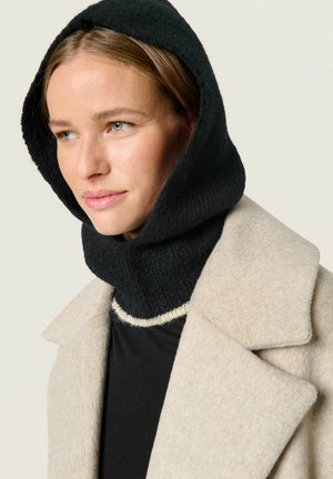 Zwarte gebreide capuchon bevestigd aan een gestructureerde beige jas. De capuchon heeft een geribbelde textuur en sluit nauw om de nek aan. Zwarte top daaronder.