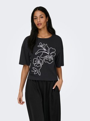 Mujer con blusa negra de corte amplio con diseño de líneas florales blancas y pantalones negros de pierna ancha, de pie con una mano en el bolsillo.