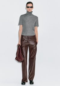 Grijze coltrui, bruine lederen wijde broek en zwarte klompen. Model houdt een bordeauxrode tas vast, achtergrond is effen wit.